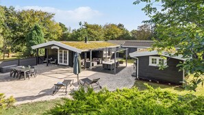 A covered outdoor patio area with seating and tables, surrounded by greenery and a small building.