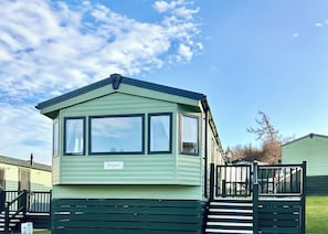A green mobile home with a nameplate reading 'Sienna,' a fenced deck, and a clear sky.