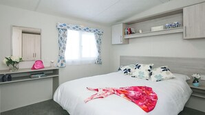 Un dormitorio con cama, tocador con espejo, ventana con cortinas, estantería con libros y un pequeño florero con flores.