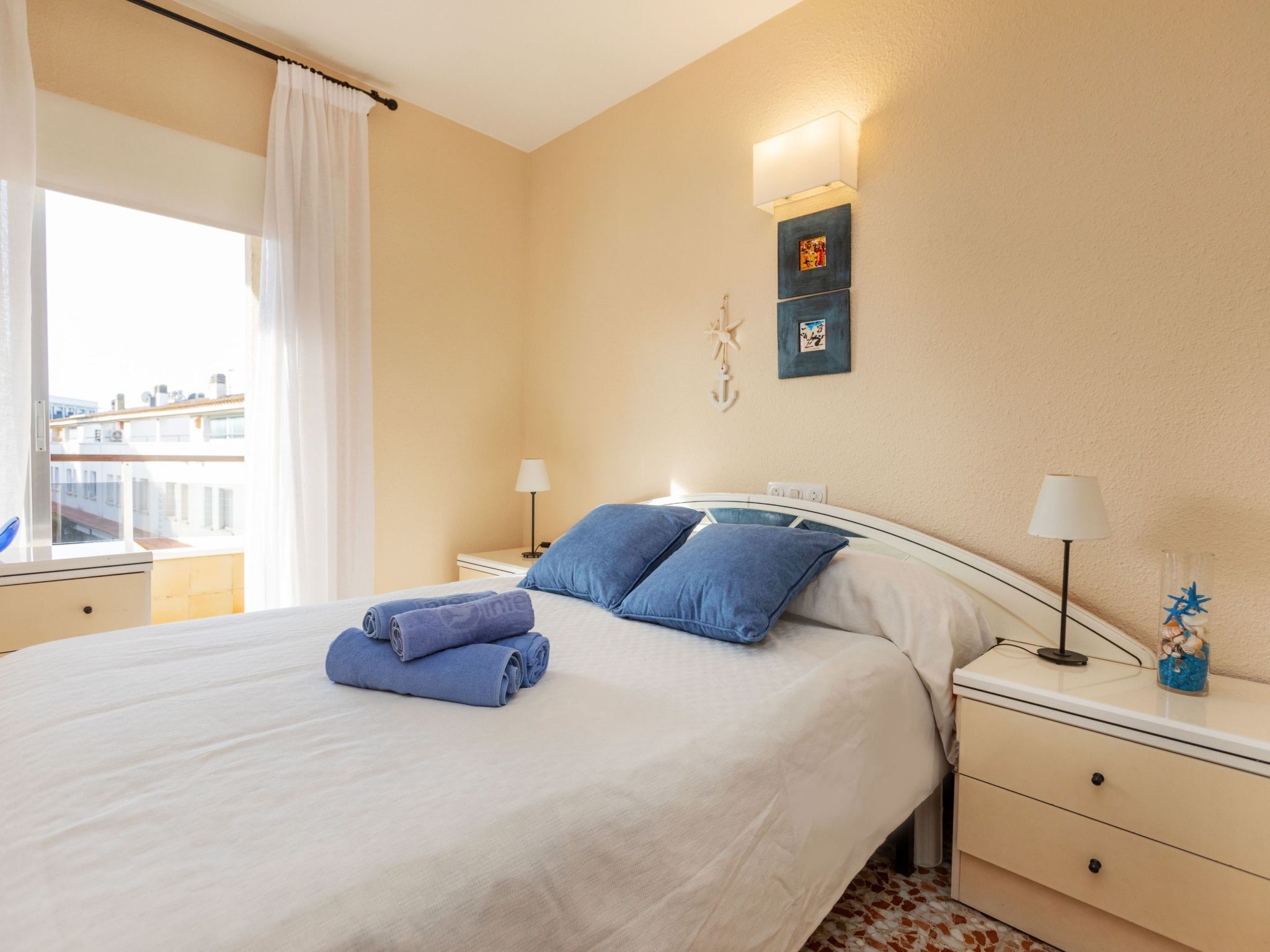 Un lit bien fait, avec des oreillers et des serviettes bleues, une table de chevet avec une lampe, et un mur orné de deux tableaux encadrés.