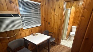 A small, wooden-paneled room with a table and two chairs, a window with blinds, and a door leading to a bathroom. - Country Cabin Motel (Quincy)