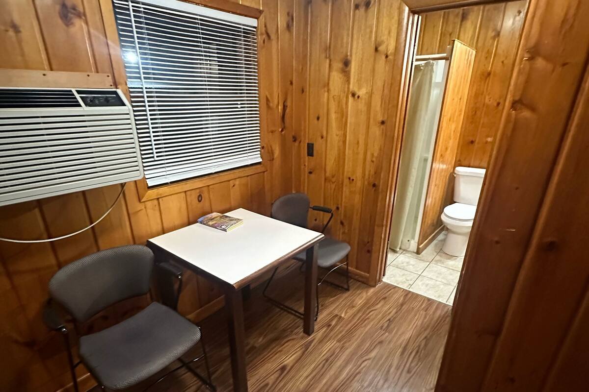 Ein kleiner Raum mit Holzwänden, einem Tisch und zwei Stühlen, einem Fenster mit Jalousien und einer Tür zu einem Badezimmer.