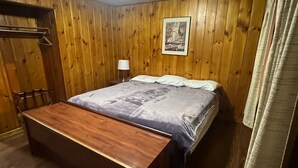 A room with wooden panel walls, a bed with a blue blanket, a wooden bench, a hanging coat rack, and a ceiling light. - Country Cabin Motel (Quincy)