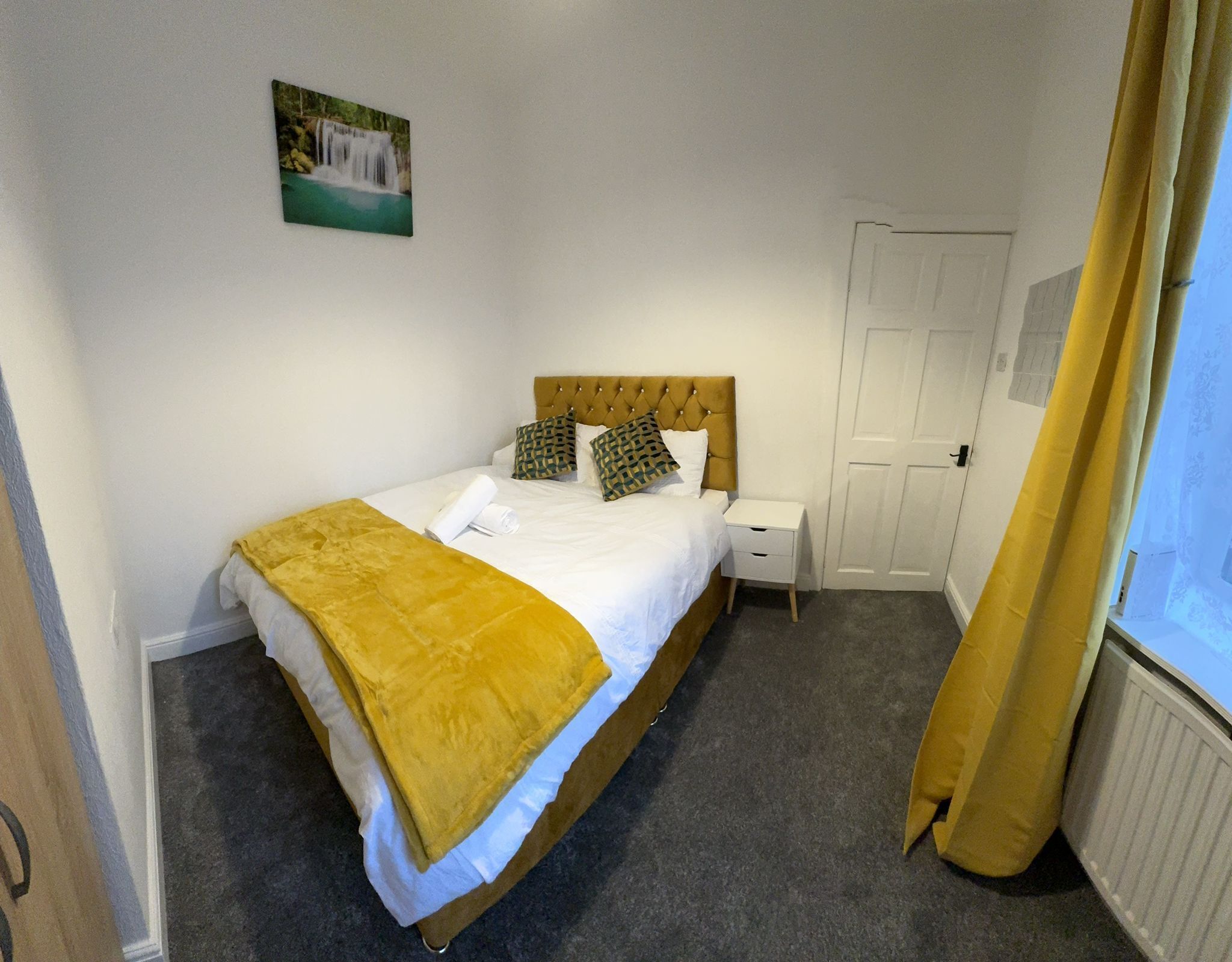 A neatly made bed with a yellow blanket, a white pillow, and a decorative pillow. There is a small bedside table with a white lamp. A door is visible in the background.