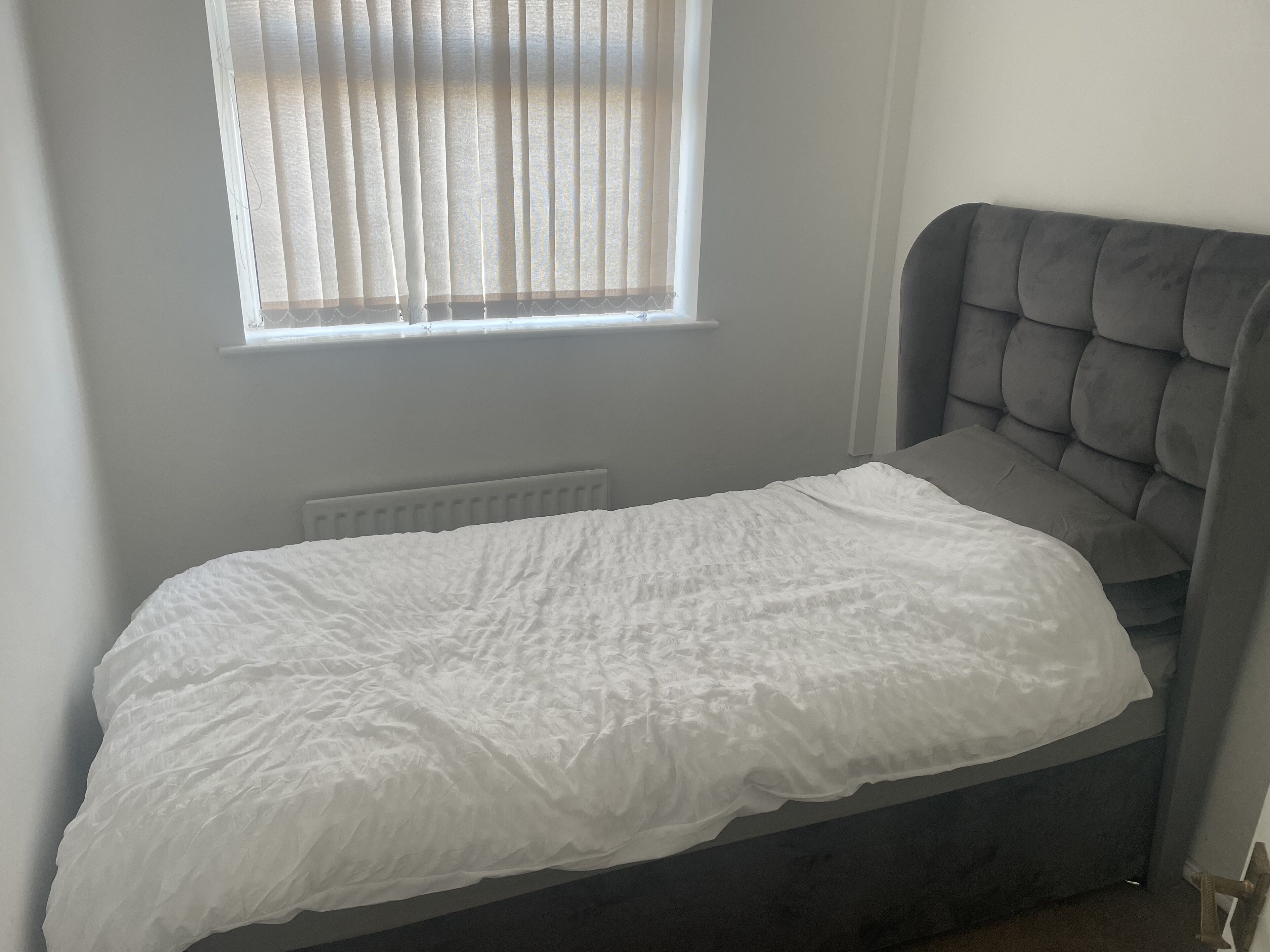 A single bed with a grey upholstered headboard, a white duvet, and a window with blinds.
