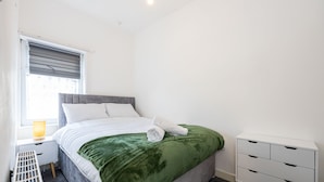 A neatly made bed with a green blanket, a bedside table with a lamp, and a white dresser. - Immaculate 3-Bed House in Accrington (Accrington)