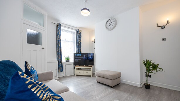 A living room with a sofa, ottoman, television, and a potted plant. - Immaculate 3-Bed House in Accrington (Accrington)
