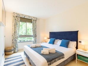Une chambre bien rangée, avec un grand lit, deux tables de chevet, une fenêtre avec des rideaux, une lampe de chevet et un petit tapis rond.