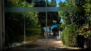 A pathway leading to a building entrance with glass doors, surrounded by greenery and a brick-paved area. - Banús, Playas del Duque, Beachfront Residential! (Puerto Banús)