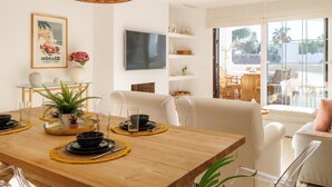A dining area with a wooden table set for four, a woven pendant light, a television on the wall, and a view of the outdoors through large windows.