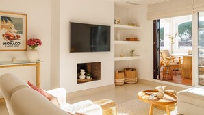 A modern living room with a flat-screen TV, a fireplace, wicker baskets, and a dining area with wicker chairs.