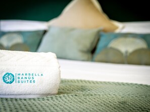 A neatly folded towel with the text 'MARBELLA BANUS SUITES' on it, placed on a bed with a green textured bedspread.