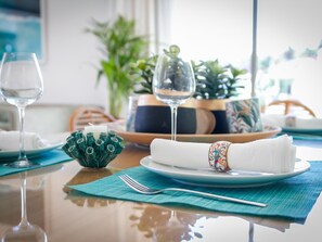 A dining table with a glass of water, a folded napkin, a plate, a fork, a decorative bowl, and a potted plant in the background.