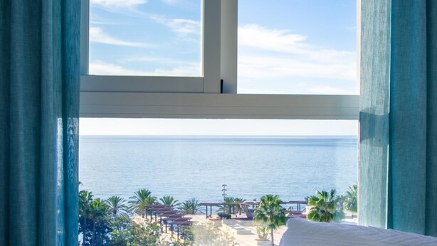 Een kamer met een groot raam met uitzicht op een strand met palmbomen en een heldere lucht.