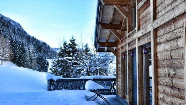 A wooden cabin with a balcony, a covered outdoor seating area, and a snow-covered landscape.