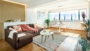 Un salon moderne avec un canapé, une table basse et un coin repas. Une grande fenêtre offre une vue sur la mer.
