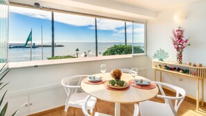 Un espace repas offrant une vue sur la mer et un pont, comprenant une table ronde entourée de chaises, un vase avec des fleurs et une étagère ornée d’objets décoratifs.