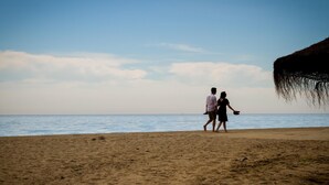A beach with two people walking towards the sea, a thatched structure on the right, and a clear sky. - Banus Playa Rocio Sea Views (Puerto Banús)