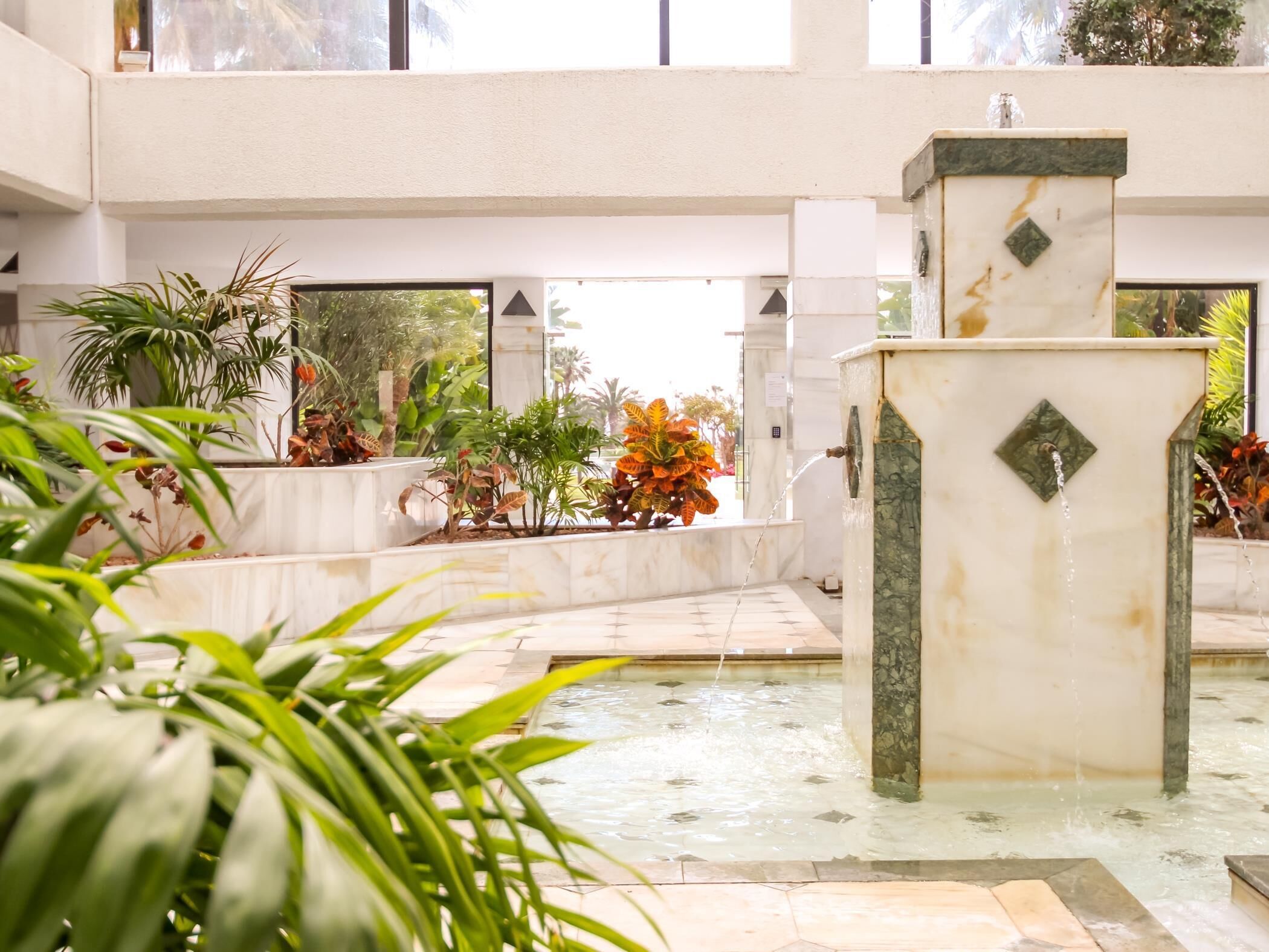 Amplo espaço interno com uma fonte central, cercado por diversos vasos de plantas e piso ladrilhado.