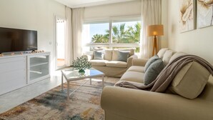 Una sala de estar moderna con un televisor de pantalla plana, una mesa de centro de vidrio, un sofá color beige y vistas a palmeras a través de amplias ventanas.