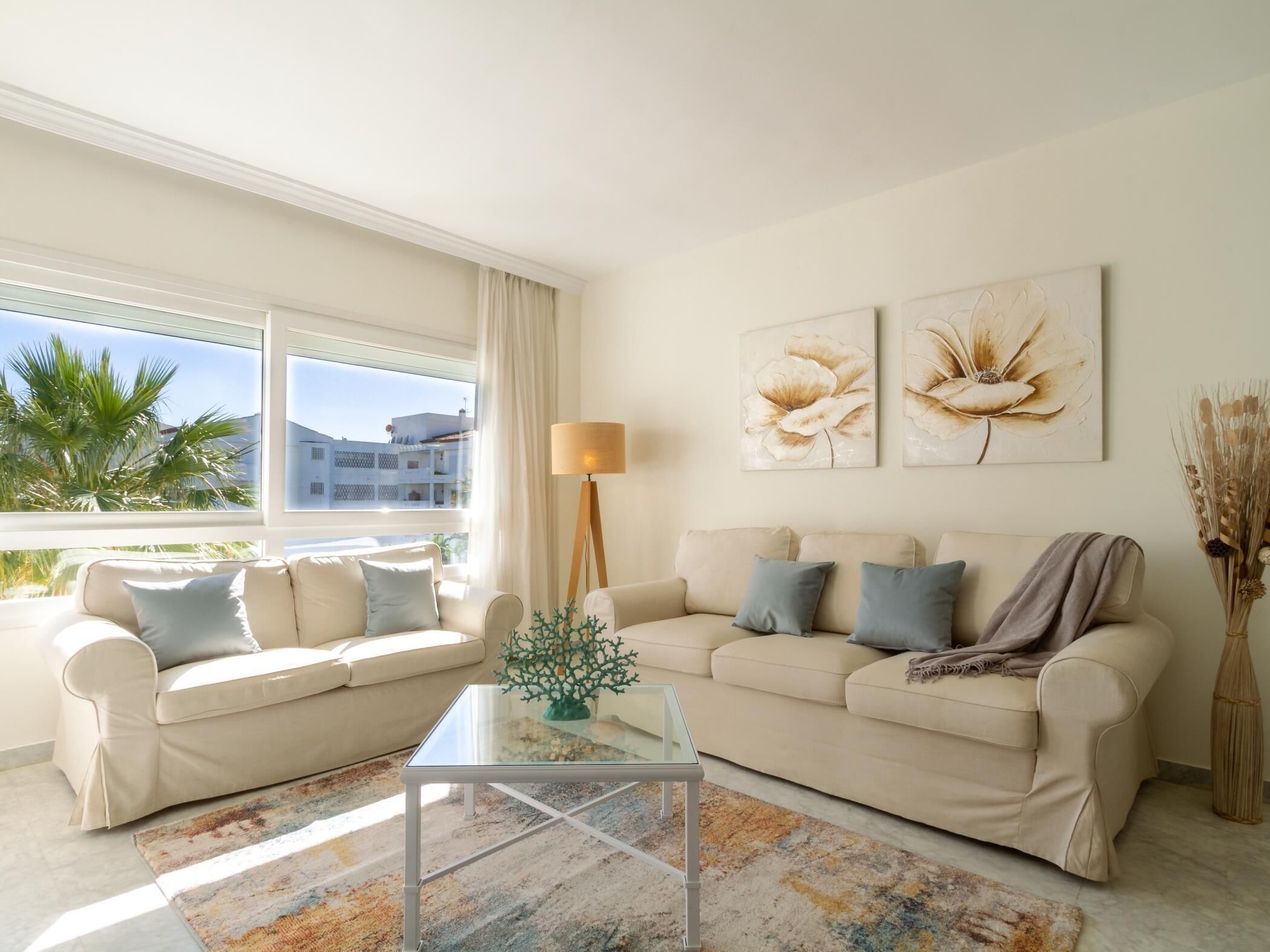 Una sala de estar con un sofá color beige, una mesa de centro de vidrio y vistas a palmeras y edificios a través de amplios ventanales.