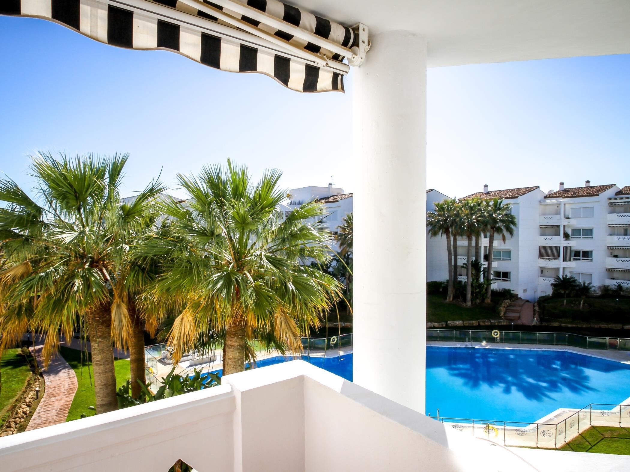 Un balcó amb vista a una piscina, palmeres i un edifici blanc amb balcons.