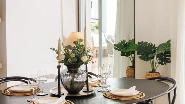 Une table à manger ornée d’un vase de fleurs, de verres à vin et d’assiettes, placée dans une pièce dotée d’un grand miroir et de plantes en pot.