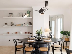 En spiseplads med et rundt bord, seks stole, en vase med blomster og et spejl på væggen.