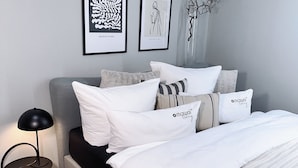A modern bedroom with a grey headboard, a bed with white and striped bedding, and two framed artworks on the wall.