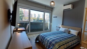 Ett sovrum med ett stort fönster, en säng med ett randigt överkast, ett sängbord, en luftkonditioneringsanläggning och en trähylla med en växt.