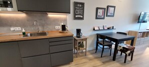 Una cocina moderna con encimera de madera, lavaplatos de acero inoxidable y cafetera. Hay una mesa de comedor con sillas y un pequeño armario con una planta.