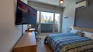 Ett modernt sovrum med en stor säng, ett skrivbord med en dator, en tv som är monterad på väggen och ett fönster med utsikt över staden.