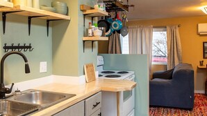 A compact kitchen with a sink, stove, and wooden cabinets. There is a dining area with a sofa and a colorful rug. - 2 bed 1 bath cottage home (Price)