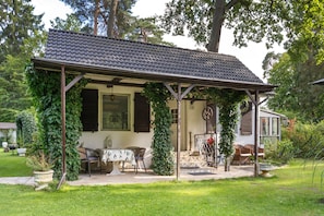 Un espace extérieur aménagé, abrité et doté d’une table et de chaises, entouré de verdure et surmonté d’un toit en tuiles.