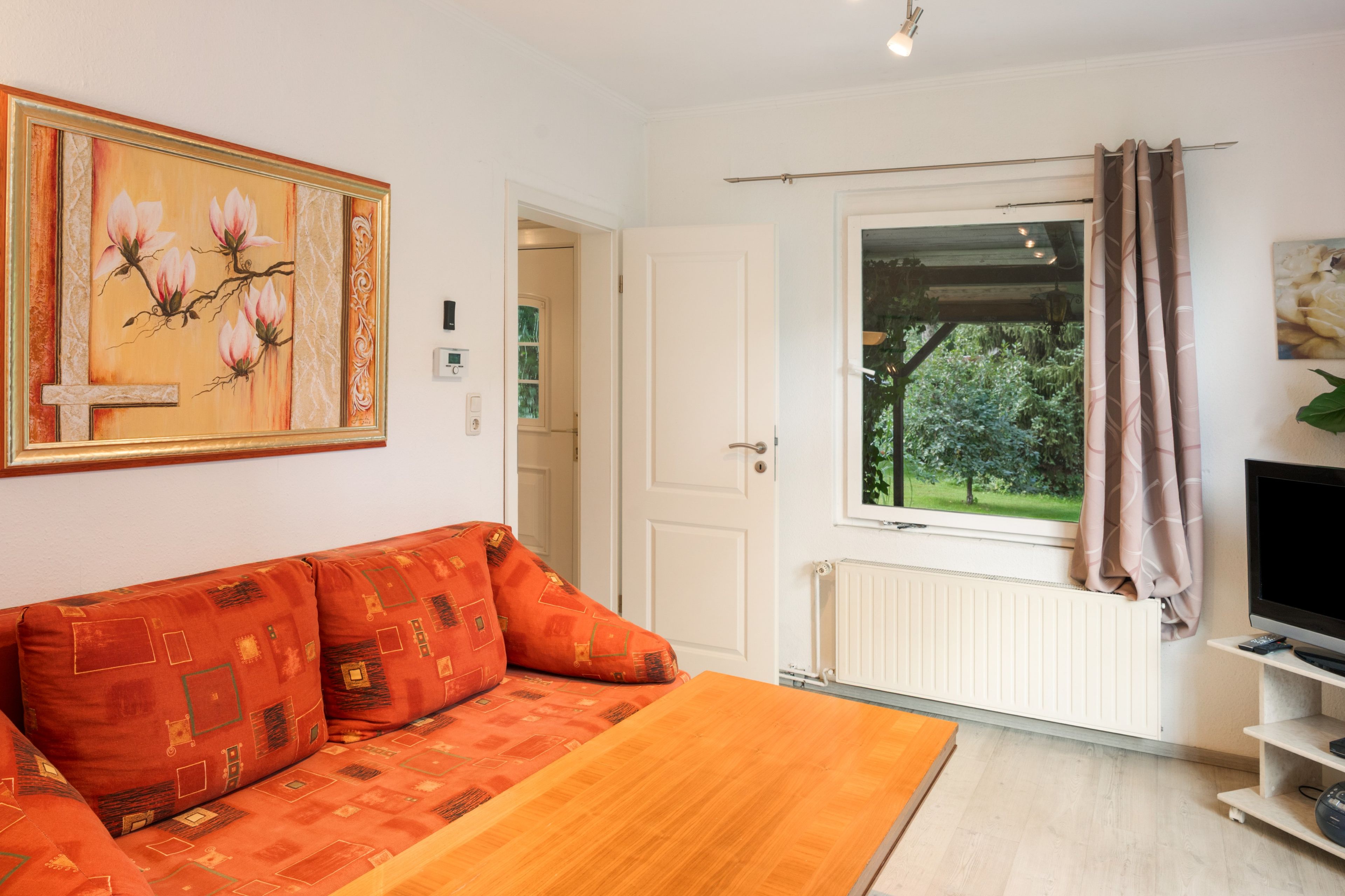 Ein Zimmer mit einer Couch, einem Fernseher, einem Fenster mit Vorhängen und einem Gemälde an der Wand.