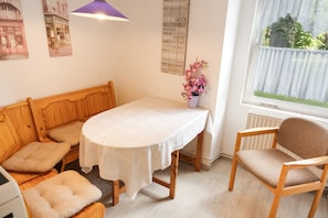 Une pièce chaleureuse avec un banc en bois, une table recouverte d’une nappe blanche, une chaise et une fenêtre donnant sur un paysage verdoyant.