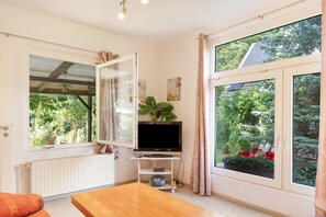 Una habitación con televisión sobre un mueble, ventana con cortinas, radiador y mesa de madera.
