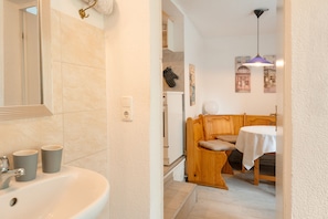 Une salle de bain avec un lavabo, un miroir et deux tasses. La pièce adjacente contient un banc en bois et une table recouverte d’une nappe.