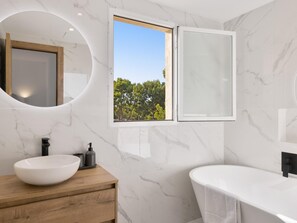 Un baño moderno con un lavamanos ovalado grande, un mueble de tocador de madera, una bañera independiente y una ventana con vistas a árboles.