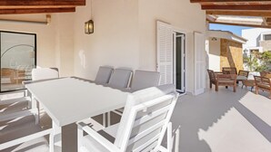 Une terrasse ensoleillée avec une table et des chaises blanches, un plafond en bois et une vue sur l’extérieur.