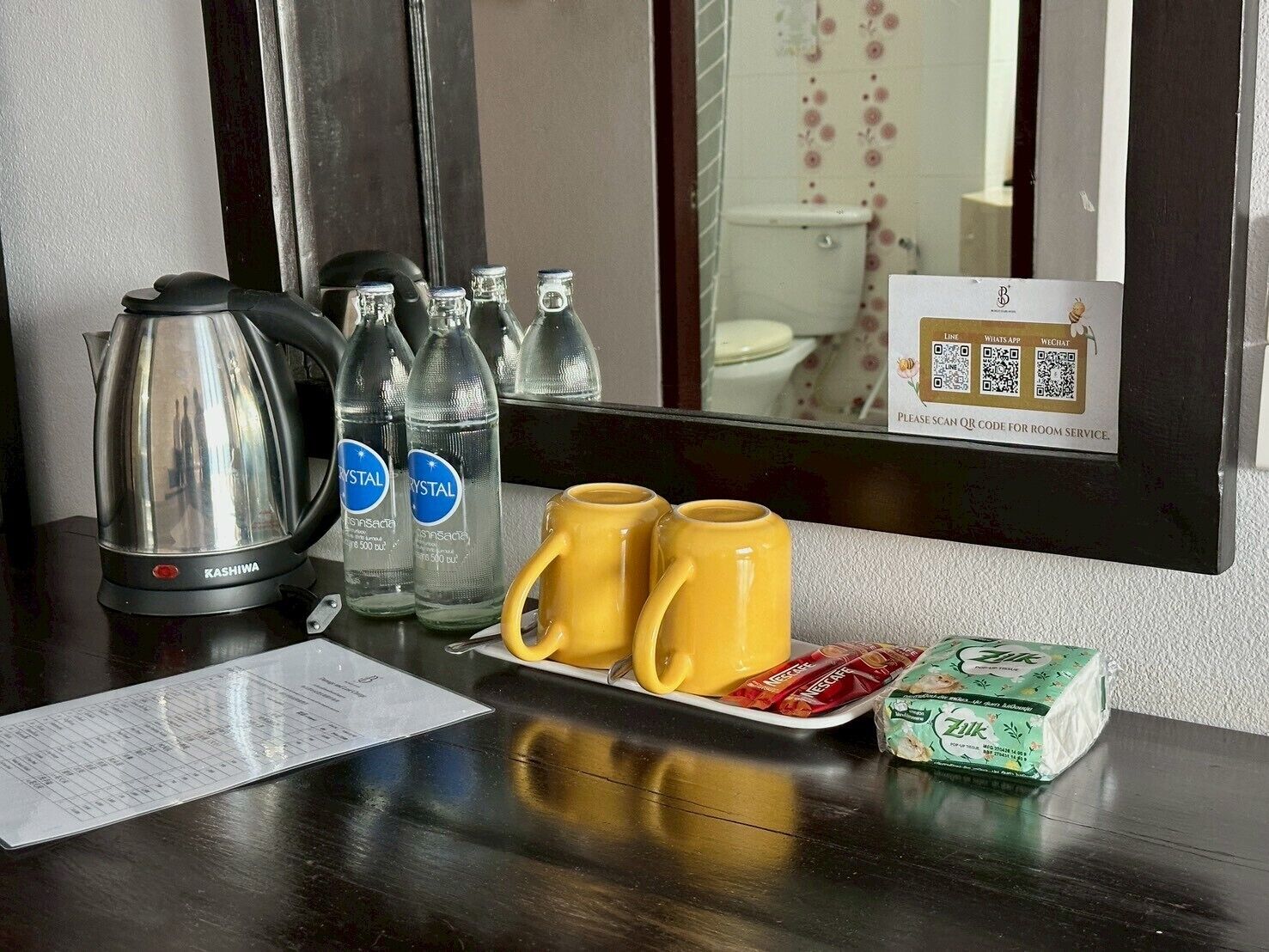 A hotel room with a coffee maker, bottled water, two yellow mugs, a tray with snacks, and a QR code sign for room service.