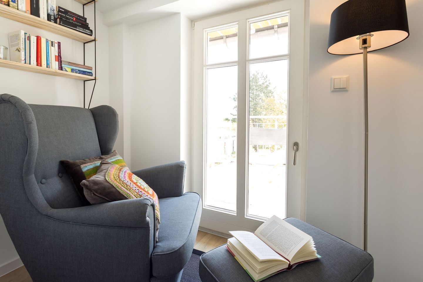 A modern living room with a grey armchair, a colorful throw pillow, a bookshelf with books, a large window with a view, and a floor lamp.