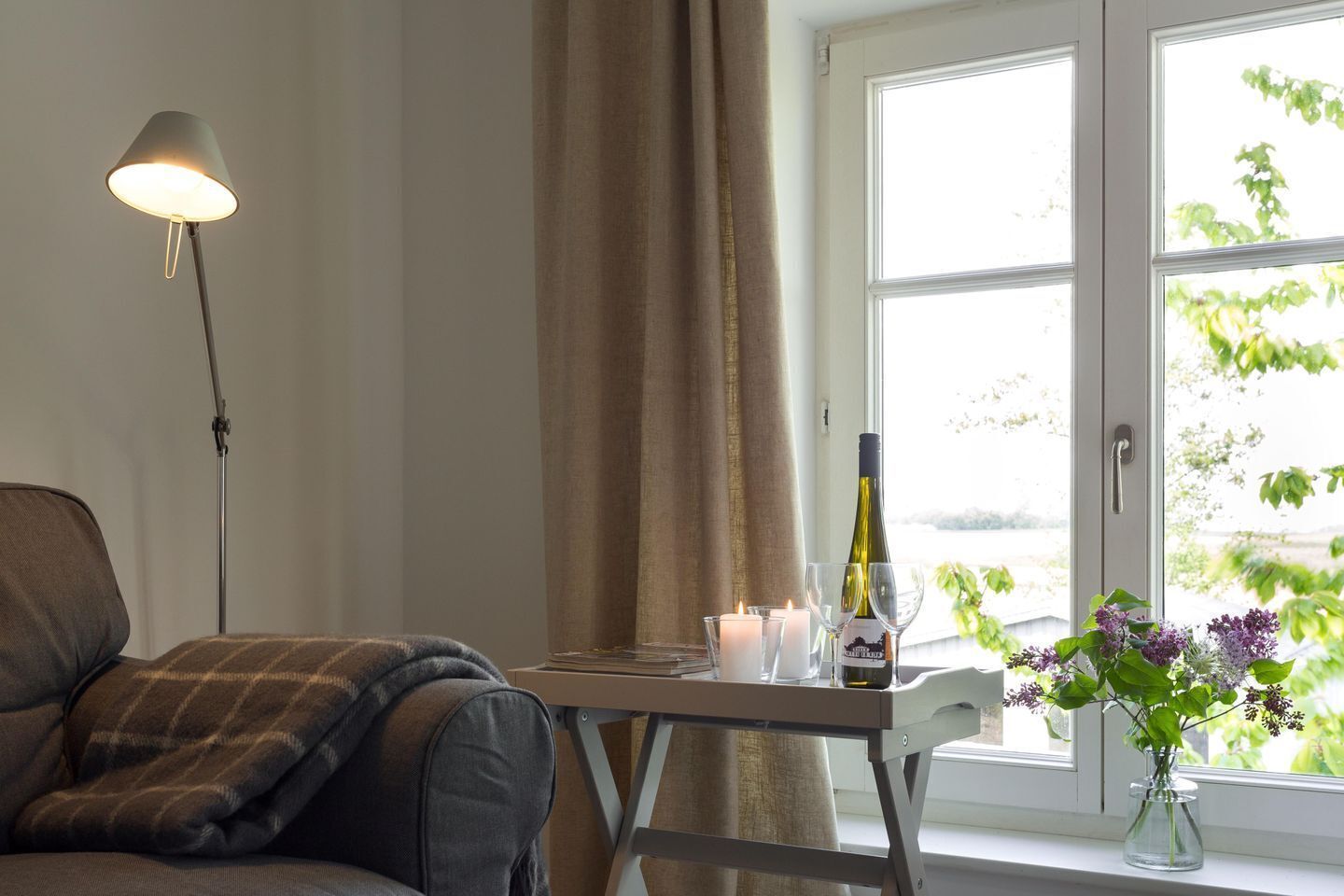 Um quarto acolhedor com um sofá, uma mesa com velas, uma garrafa de vinho e um vaso com flores perto de uma janela.