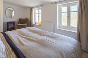 Un dormitorio con cama, una silla, una mesita, un espejo y una ventana con cortinas.