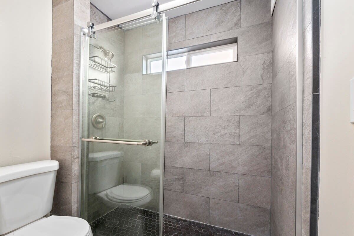 Douchecabine met glazen wanden, een toilet links, een handdoekhouder en een raam boven het toilet.