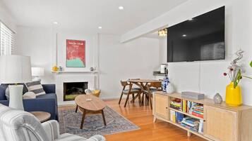 Una sala de estar moderna con chimenea, un televisor de pantalla plana montado en la pared, un comedor con mesa y sillas de madera, y una estantería llena de libros.