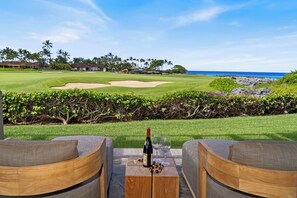 A golf course with a view of the ocean, a wooden table with a bottle and glasses, and two wooden chairs.