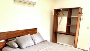 Een slaapkamer met een houten bed, grijze beddengoed, een houten garderobe met planken, en een wandairconditioner.