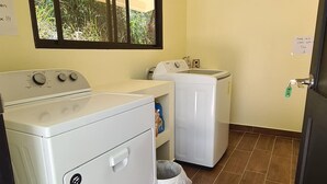 Een wasserette met een wasmachine, droger en een raam met uitzicht op groen.