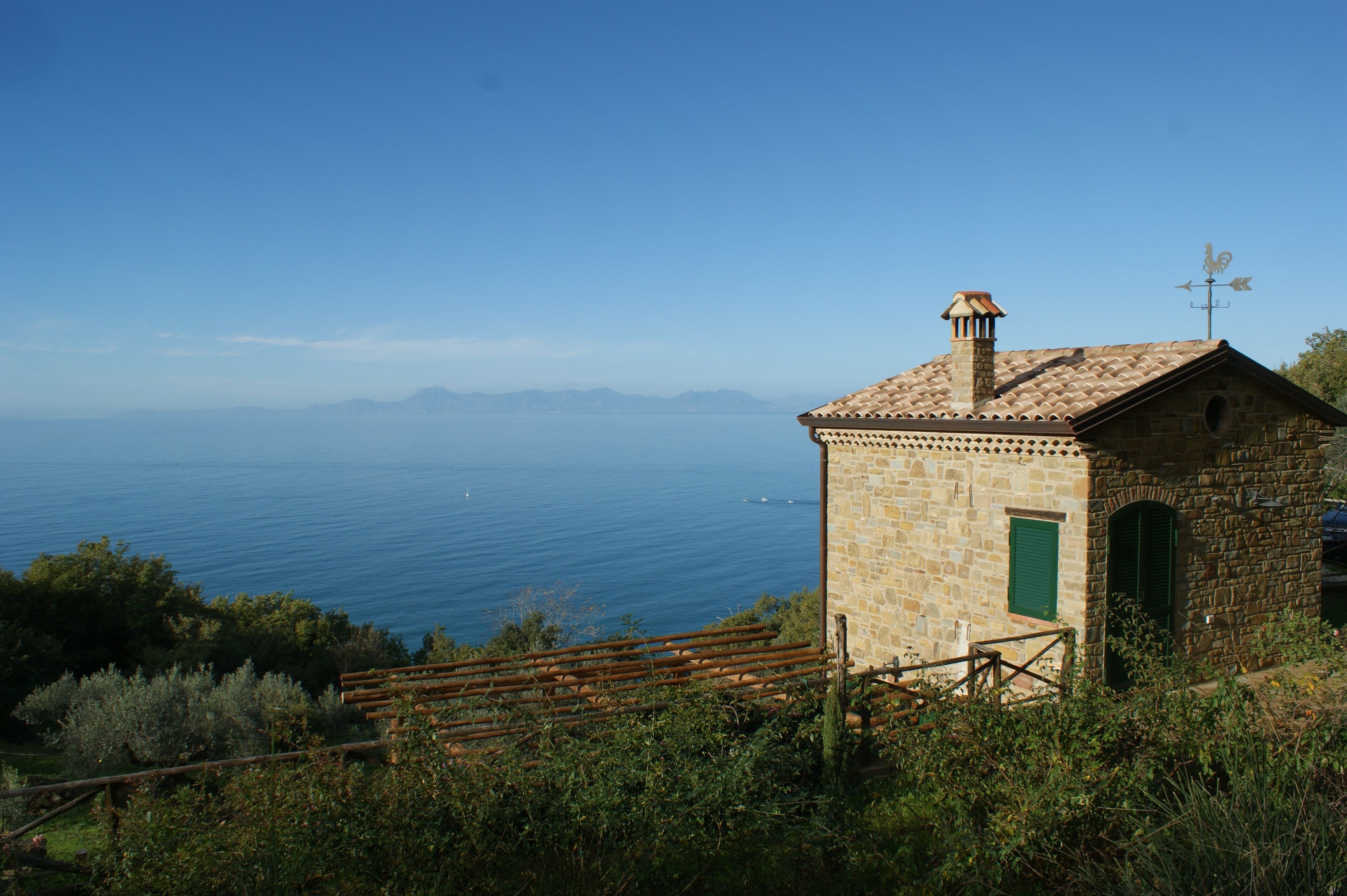 A stone building with a tiled roof, a green door, and a small window. It is surrounded by greenery and overlooks a body of water.